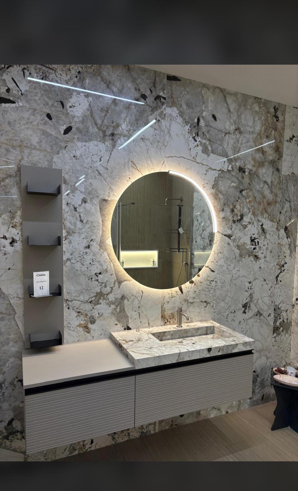 Top con lavabo integrato in gres effetto marmo, dello stesso materiale della parete, in uno showroom con specchio tondo retroilluminato e mensole a giorno
