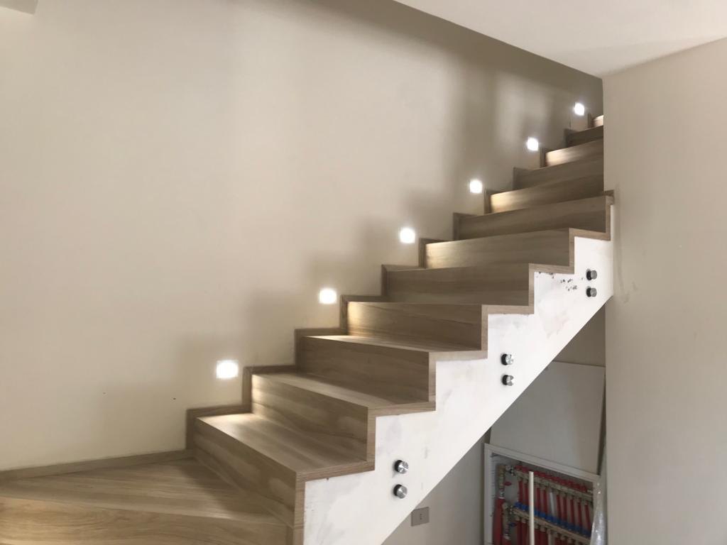 Scala a rampa unica rivestita con gradini in gres effetto legno chiaro e luci segnapasso a parete, con il fianco ancora da rifinire