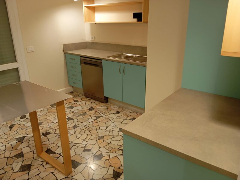 Cucina con piano e alzata in gres effetto cemento, lavello incassato nel top e mobili color verde acqua; a terra un pavimento in palladiana di marmo